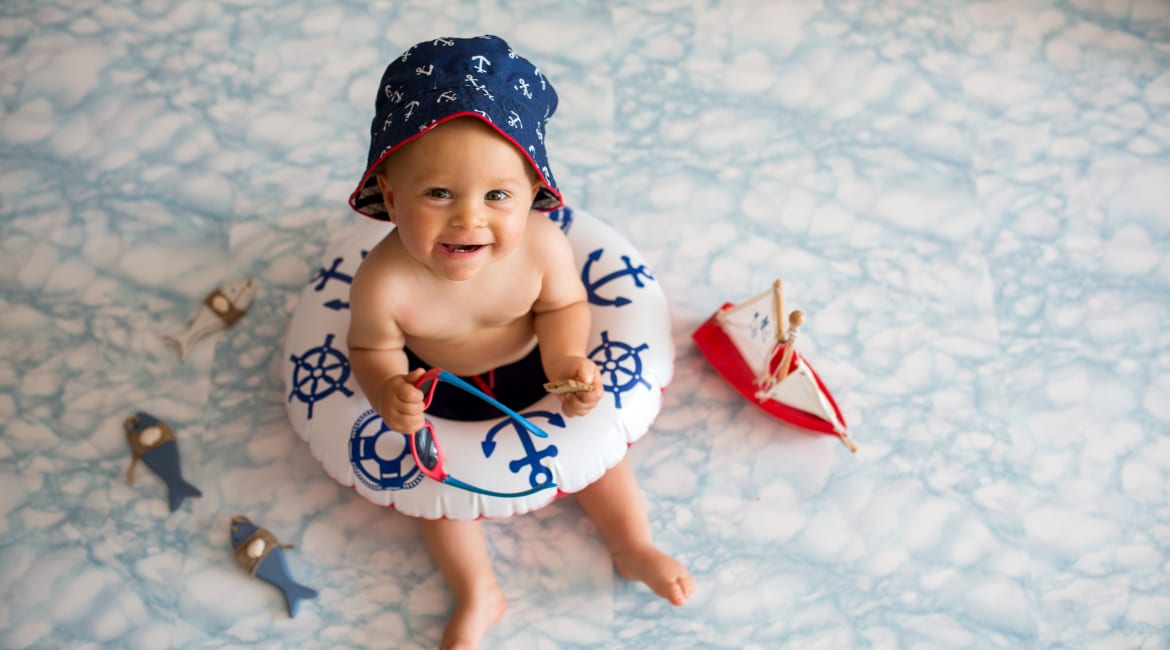 Cute baby boy lying down on a tiny inflatable swim ring, wearing swimsuit shorts and sunglasses, indoor shot