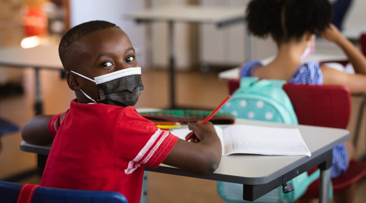 Portrait of african american boy wearing face mask sitting on his desk in class at elementary school