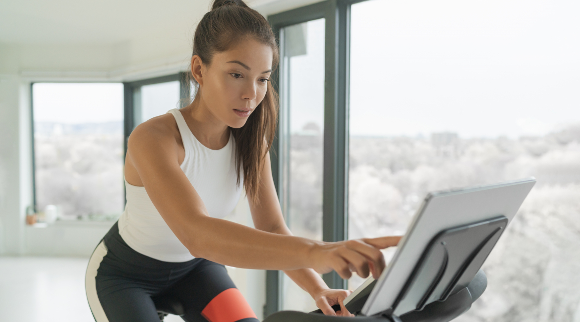 Home cycle workout Asian fit athlete woman training on stationary bike doing online cycling exercise class biking during livestream on screen.