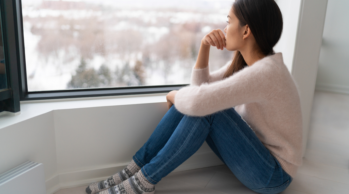Depressed young girl feeling sad an lonely, anxious looking out the window in winter. Unhappy Asian woman alone, depression at home.